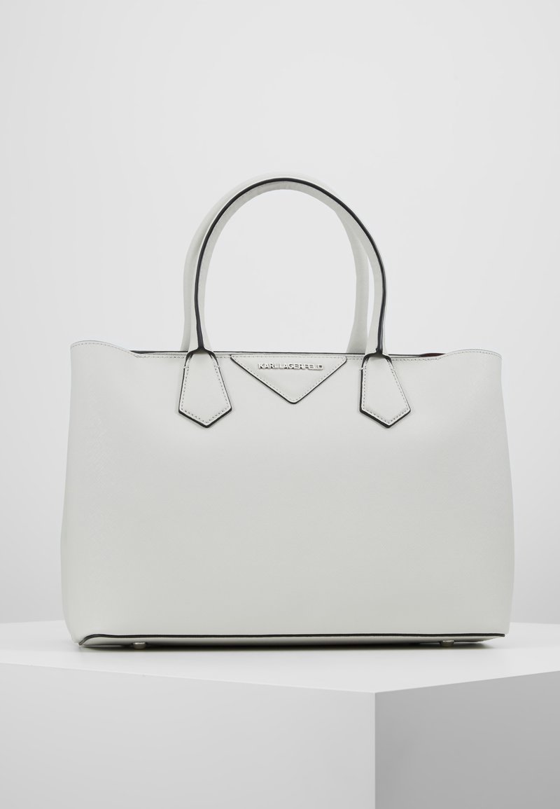 Sac cabas en cuir blanc avec bordure noire, double poignée et une petite plaque triangulaire avec logo à l'avant.