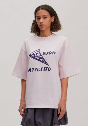 Vrouw met krullend haar draagt een oversized lichtroze shirt met een pizzapunt en de tekst "Buon Appetito" in donkerblauw.