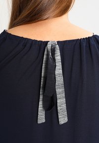 Laivastonsininen yläosa joustavalla kaula-aukolla ja solmittavalla suljennalla. Solmussa on harmaa raidallinen yksityiskohta. Sileä koostumus ja rento istuvuus.