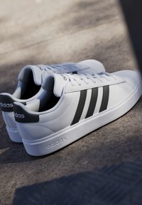 Paar weiße Adidas-Sneaker mit schwarzen Streifen und Logo, auf einer strukturierten Außenfläche im natürlichen Licht platziert.