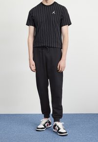 Černé pruhované tričko s malým logem, černé kalhoty se šňůrkou a černobílé červené tenisky na modrém pozadí.