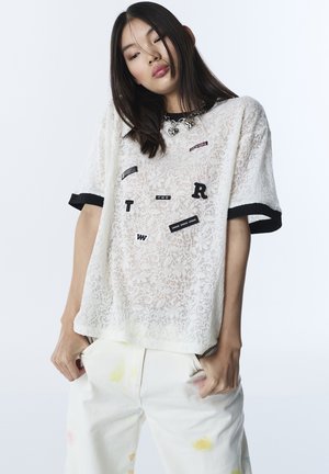 Giovane donna con lunghi capelli scuri che indossa una camicia bianca di pizzo con lettere nere e pantaloni bianchi con lievi macchie gialle e rosse.