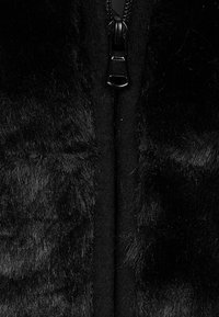 Schwarzer Kunstpelzstoff mit glatter Textur und einem zentralen schwarzen Reißverschluss. Das Material wirkt plüschig und weich und verstärkt seine optische Tiefe.