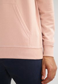 Felpa rosa chiaro con tasca frontale, polsini e orlo a coste. Il tessuto sembra morbido, con dettagli di cucitura visibili e una vestibilità rilassata.