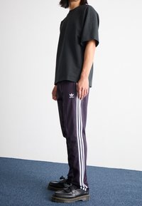 Person som bär en svart kortärmad skjorta, lila Adidas träningsbyxor med vita ränder och svarta platåskor, stående på en blå matta.