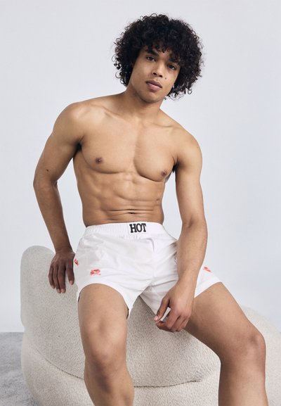 Pantaloni corti bianchi con "HOT" stampato sulla vita, con accenti rossi. Il modello ha una corporatura muscolosa e capelli ricci, seduto su una sedia strutturata.