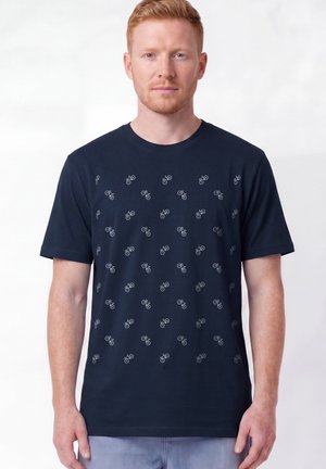 Mann mit roten Haaren, der ein dunkelblaues T-Shirt mit kleinen weißen Fahrradmustern trägt und vor einem schlichten hellen Hintergrund steht.