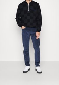 Pullover a scacchi neri con colletto con zip, abbinato a pantaloni blu navy. Calzature: scarpe bianche con dettagli neri.