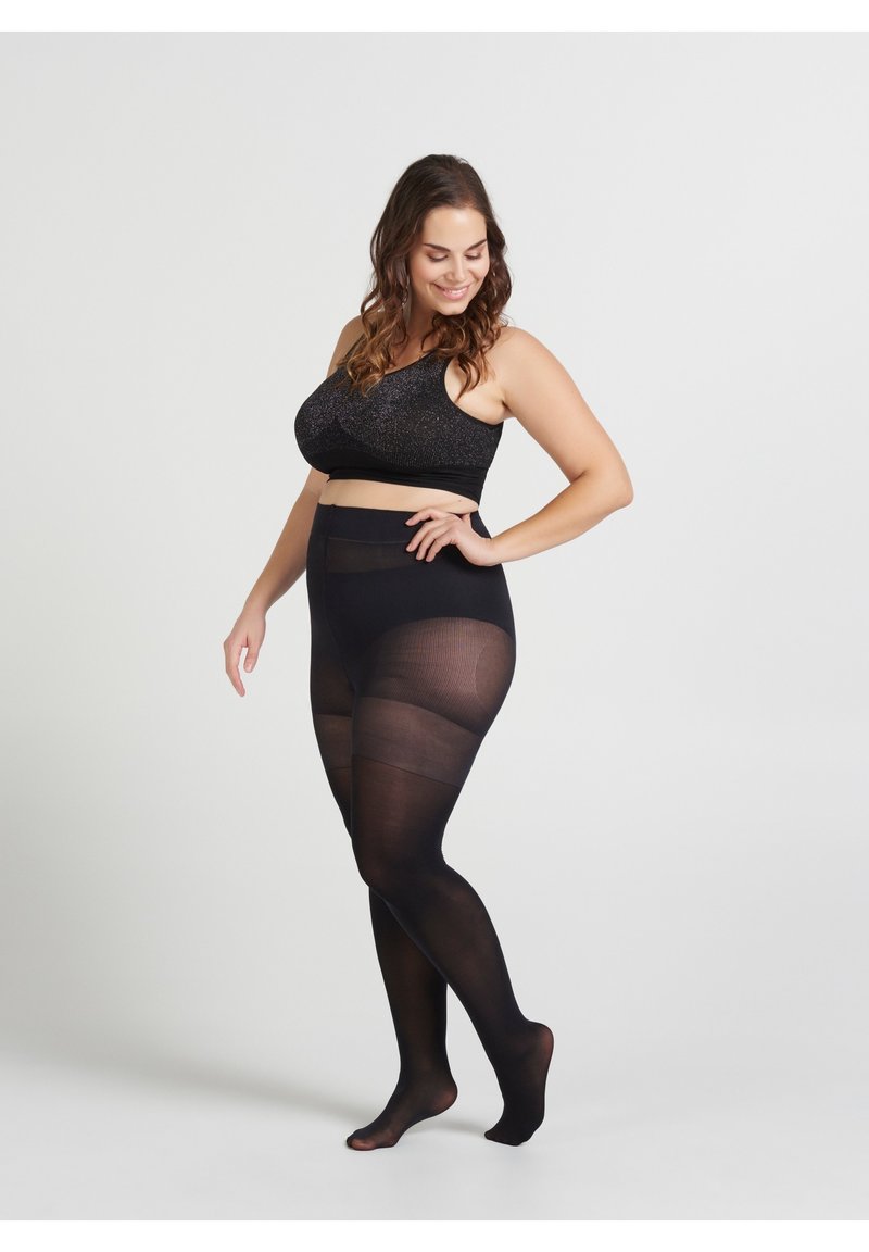 Donna curvilinea che posa a piedi nudi, indossando un crop top nero scintillante e collant neri trasparenti su uno sfondo bianco di uno studio.