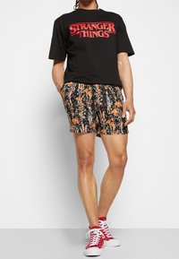 T-shirt noir avec le texte "STRANGER THINGS" en rouge, associée à un short à imprimé floral en noir, orange et blanc, et des sneakers montantes rouges.