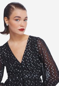 Abito nero con scollo a V profondo e maniche lunghe trasparenti, con motivi quadrati argentati e una vita con fiocco, tessuto testurizzato. Orecchini a forma di cuore.