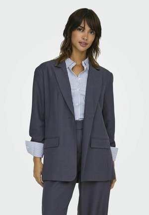 Kvinde iført en oversized mørkegrå blazer og stribet skjorte med knapper samt matchende grå bukser mod en ensfarvet hvid baggrund.