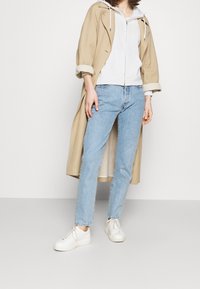 Beige trench coat over a light grey zip-up hoodie, blue jeans, and white sneakers. Casual and relaxed fit with rolled sleeves.