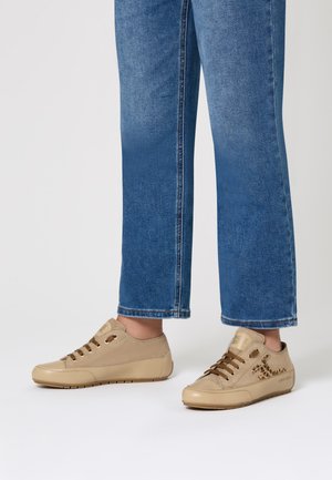 Persona che indossa jeans blu e sneakers beige con dettagli a stampa leopardata e lacci marroni, in piedi su una superficie bianca.