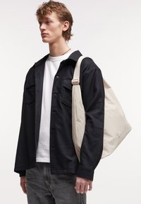 Sac fourre-tout beige avec une sangle adjustable, en matériau à texture lisse et avec un logo discret. Porté par une personne en veste noire et chemise blanche.