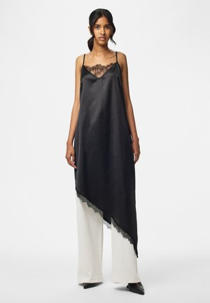 Femme portant une robe nuisette noire asymétrique avec un bord en dentelle sur un pantalon blanc à jambes larges et des chaussures noires pointues, debout devant un fond uni.