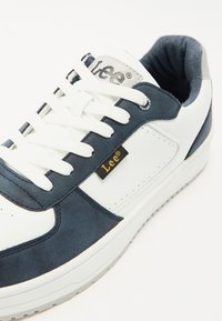 Zapatillas blancas y azul marino que combinan materiales de cuero y ante, con punta redondeada, cordones gruesos blancos y una etiqueta de marca negra y dorada.