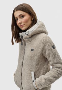 Veste en polaire beige avec capuche, fermeture éclair et accents gris clair contrastés. Comprend des poches, une surface texturée et des détails de logo.