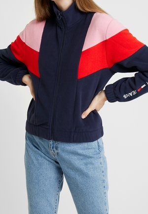 Fleece zip-up jack met een marineblauwe basis en kleurblokken in roze, rood en wit. Zachte textuur en ribgebreide manchetten aanwezig.