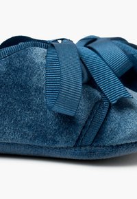 Pantofole in velluto blu caratterizzate da una forma arrotondata, una consistenza morbida e nastri in satin decorativi. Il materiale appare morbido e invitante.