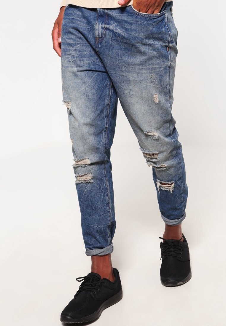 Homme portant un jean bleu clair usé avec les revers roulés et des baskets noires, debout avec une main dans la poche.