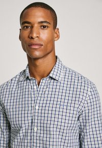 Camisa de cuadros en blanco y azul marino, con cuello, mangas largas y un pequeño logo en el pecho. Textura de tela suave.