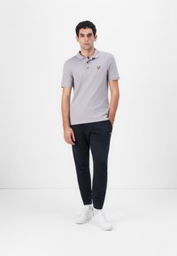 Grå polo skjorte med en gul logo på brystet, kombinert med mørkeblå joggebukser og hvite sneakers. Modellen står mot en enkel bakgrunn.