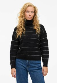 Črna turtleneck pulover s horizontalnimi srebrnimi črtami, rebrasto teksturo in ohlapnim krojem. Kombiniran z modrimi kavbojkami za sproščen videz.