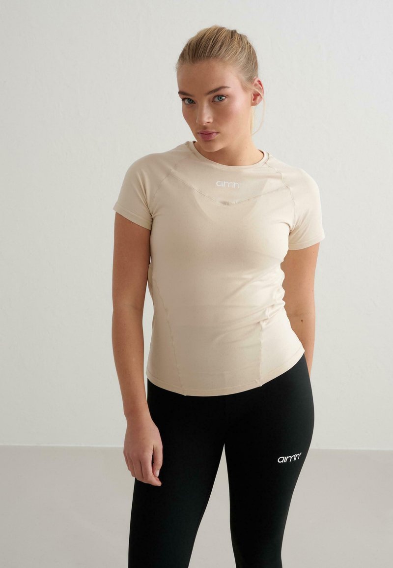 Beiges, tailliertes Kurzarmshirt mit rundem Ausschnitt und dezentem Logo auf der Brust, kombiniert mit schwarzen Leggings, die ein weißes Logo tragen.