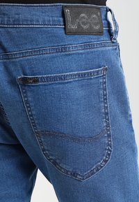 Blå denimjeans med klassisk skärning, en svart läderlogotyp på midjebandet och en enda bakficka med sydda detaljer.