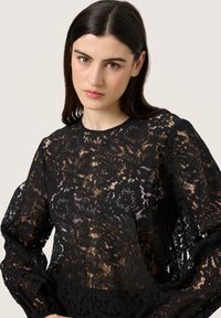 Blusa nera in pizzo con motivo floreale, maniche lunghe e scollatura rotonda. Il tessuto trasparente rivela la pelle sottostante, creando un effetto a strati.