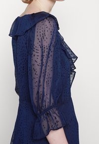 Navy sheer blouse dengan detail ruffle dan kain bertekstur. Memiliki lengan puff yang berakhir dengan manset elastis, dengan pola bercak halus.