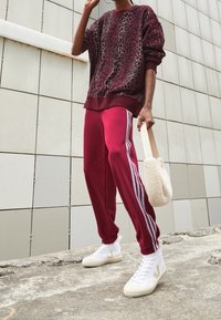 Burgundy leopardmönstrad sweatshirt, matchande träningsbyxor med vita ränder, och vita högklackade sneakers. Håller en krämfärgad väska.