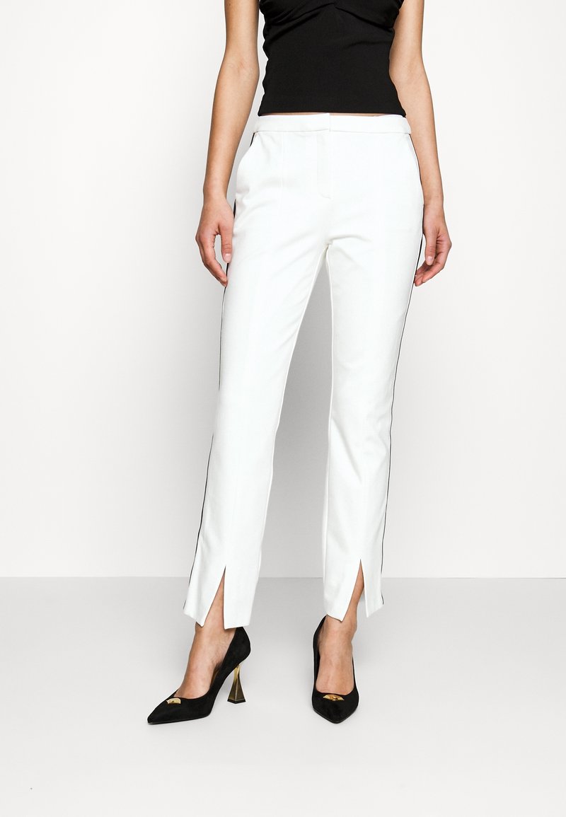 Femme portant un pantalon blanc slim avec des fentes à l'avant et des talons hauts noirs, associé à un haut noir sans manches sur un fond uni.