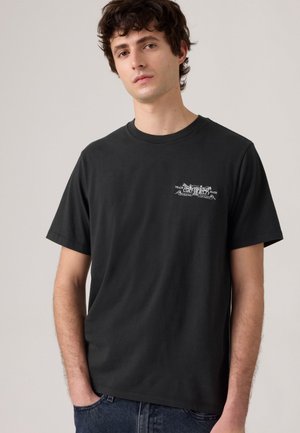 T-shirt en coton noir à manches courtes, col rond, avec un petit imprimé graphique blanc sur le côté gauche de la poitrine, présentant un design simple.