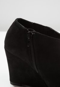 Black wedge ankle boot made of soft suede, featuring a side zipper and elastic panel for easy wear. Smooth texture and solid color.