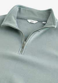 Pullover verde chiaro a maniche lunghe con colletto a zip e texture liscia. Presenta una cerniera argentata e un'etichetta che indica la taglia M.
