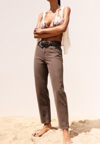 Jean marron taille haute avec jambes droites, assorti à un haut en crochet blanc et une ceinture tressée noire, sur un fond sablonneux.