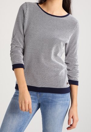 Kobieta w teksturowanej granatowo-białej bluzce z długim rękawem i granatowymi wykończeniami, ubrana w jasnoniebieskie dżinsy, stojąca na jednolitym tle.