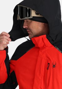 Rote und schwarze isolierte Jacke mit Kapuze, die ein Logo auf der Brust, verstellbare Kordelzüge und eine Reißverschlusstasche aufweist. Texturiert.