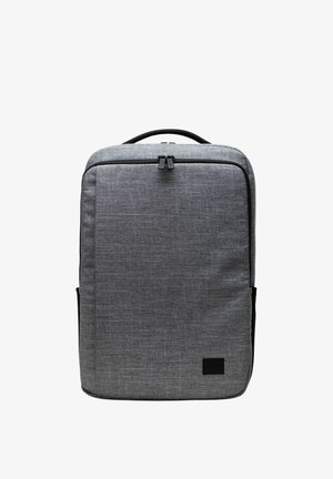 Sac à dos rectangulaire en tissu gris avec poignée supérieure, compartiment avant zippé et patch logo noir dans le coin inférieur droit.