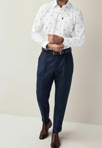 Camisa blanca de manga larga con pequeños detalles en negro y verde, combinada con pantalones oscuros de textura azul y zapatos de vestir marrones.