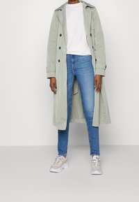 Persona che indossa un trench lungo grigio chiaro sopra una camicia bianca, jeans skinny blu e sneakers beige, in piedi contro uno sfondo semplice.