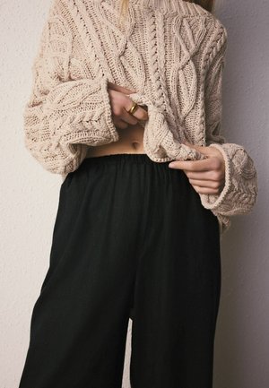 Persoană ridicând un pulover bej cu model împletit pentru a dezvălui abdomenul gol, purtând pantaloni negri și un inel de aur la mâna stângă.