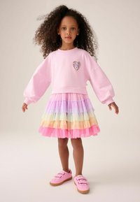 Ragazza con capelli ricci che indossa una felpa rosa con una toppa a forma di cuore, una gonna di tulle a strati con colori pastello arcobaleno e scarpe da ginnastica rosa, in piedi con le braccia aperte.