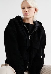 Černá fleecová mikina s předním zipem, dvěma kapsami na hrudi a stahovacími šňůrkami. Má texturovanou látku a casual design.