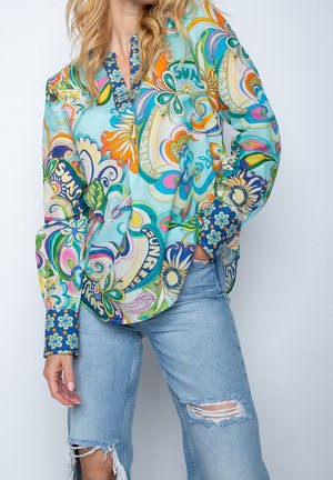 Kvinde iført en langærmet bluse med farverige mønstre, blomster- og abstrakte designs, kombineret med lyse, revnede jeans.