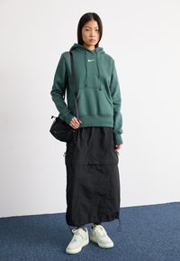 Groene katoenen hoodie met een voorzak en wit logo, gecombineerd met een zwarte maxi rok en mintgroene sneakers. Zwarte crossbody tas.
