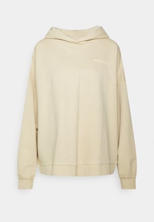Beżowa, oversize bluza z kapturem z długimi rękawami, ściągaczami przy rękawach i dole oraz małym białym logo na górnej lewej części klatki piersiowej.