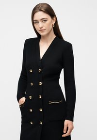 Γυναίκα που φοράει εφαρμοστό μαύρο ζακέτα με χρυσά κουμπιά και αλυσίδες στα τσέπες, ποζάρει με το ένα χέρι στην τσέπη μπροστά σε απλό φόντο.